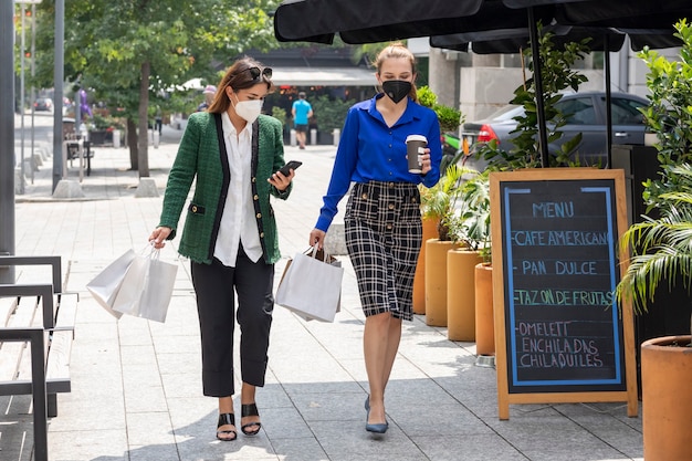 Two young women are walking down the street wearing face masks talking happily after shopping