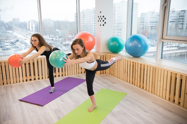 Two young women balancing on one leg holding a ball in the fitness studio