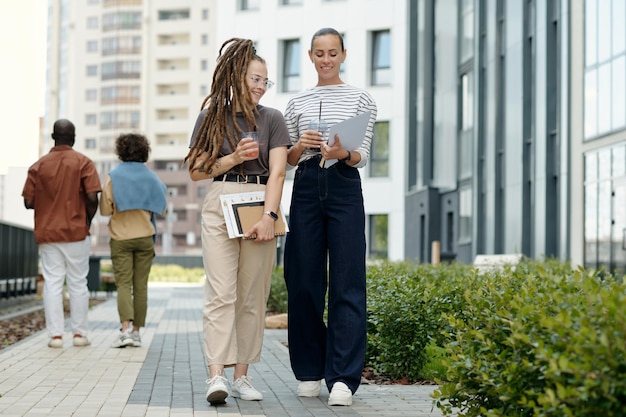 Two young women in casualwear discussing papers while walking to office