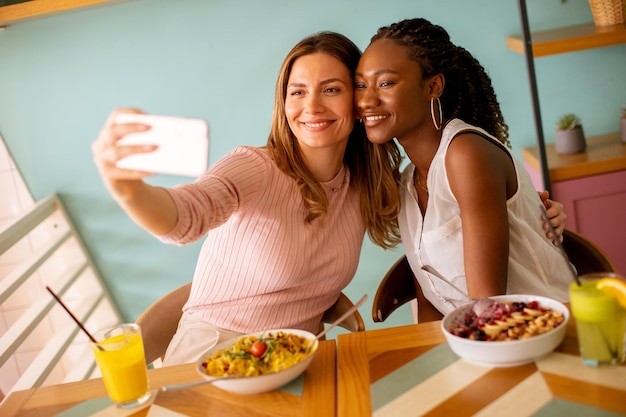 2 人の若い女性白人と黒人の 1 つは、カフェで携帯電話で selfie を取っています。