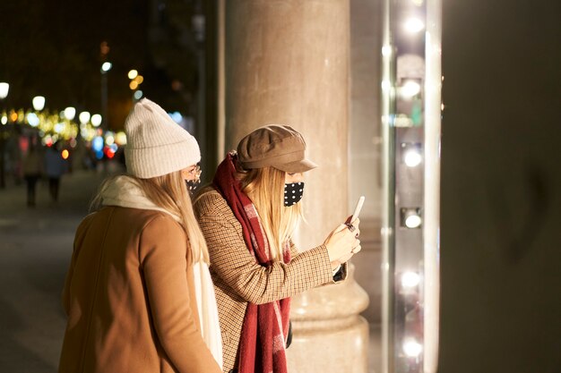 夜の街で冬服を着て店の窓を見ている2人の若い女性。そのうちの1人は彼女の電話で何かの写真を撮ります。