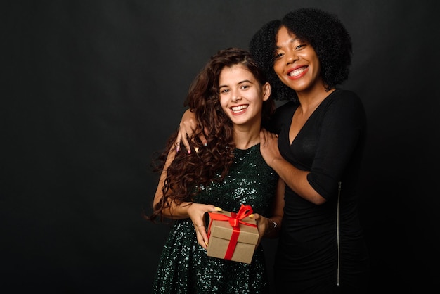 Two young women of different nationalities are hugging holding a gift for the new year and christmas