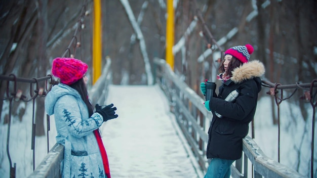 Two young women drinking hot drinks from the thermos and walk on the bridge