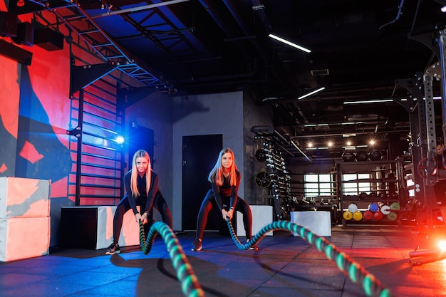 Two young women exercising with ropes ropes in the gym. Woman doing rope workout together with trainer