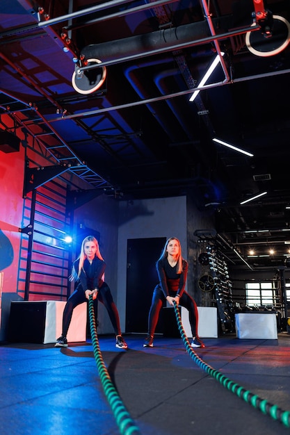 Two young women exercising with ropes ropes in the gym. Woman doing rope workout together with trainer