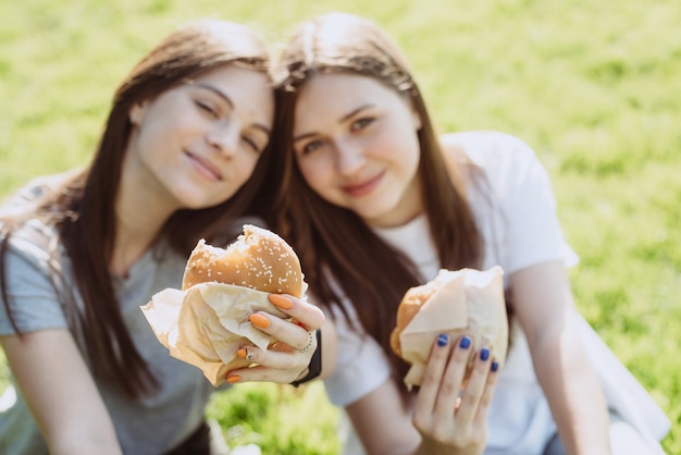 Две молодые женщины друзья едят вкусные гамбургеры в парке на траве. не здоровая диета. мягкий выборочный фокус, расфокусировка.