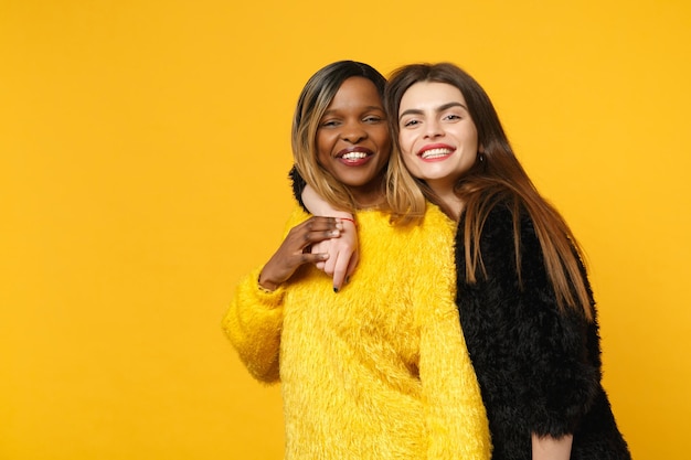 Two young women friends european and african american in black yellow clothes standing posing isolated on bright orange wall background, studio portrait. People lifestyle concept. Mock up copy space.