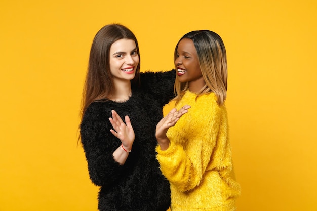 Photo two young women friends european and african american in black yellow clothes standing posing isolated on bright orange wall background, studio portrait. people lifestyle concept. mock up copy space.