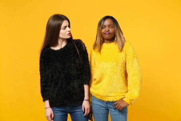 Two young women friends european and african american in black yellow clothes standing posing isolated on bright orange wall background, studio portrait. People lifestyle concept. Mock up copy space.