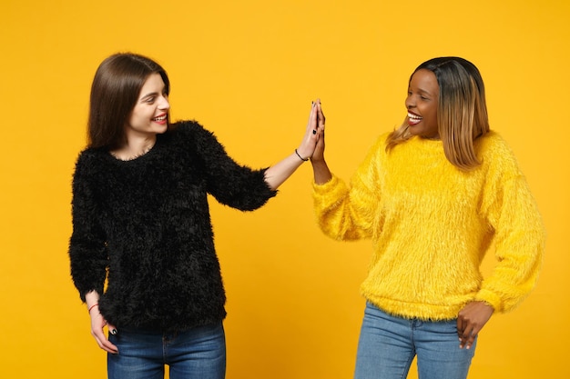 Two young women friends european and african american in black yellow clothes standing posing isolated on bright orange wall background, studio portrait. People lifestyle concept. Mock up copy space.