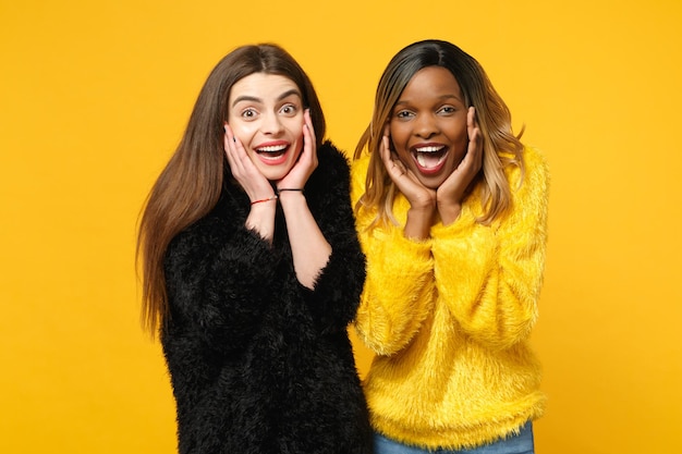 Two young women friends european and african american in black yellow clothes standing posing isolated on bright orange wall background, studio portrait. People lifestyle concept. Mock up copy space.