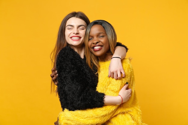 Two young women friends european and african american in black yellow clothes standing posing isolated on bright orange wall background, studio portrait. People lifestyle concept. Mock up copy space.