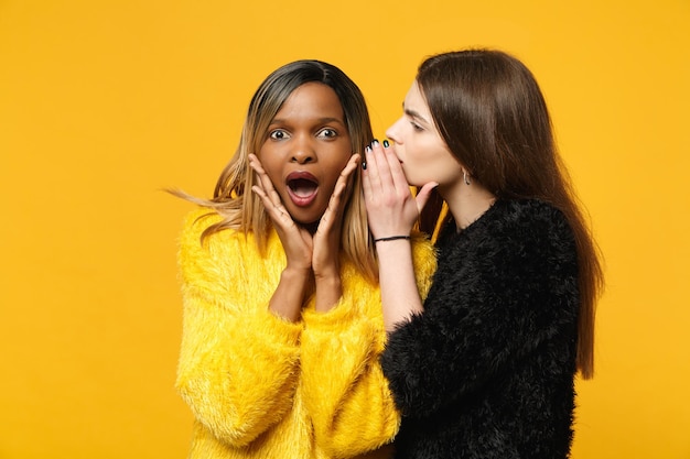 Two young women friends european and african american in black\
yellow clothes standing posing isolated on bright orange wall\
background, studio portrait. people lifestyle concept. mock up copy\
space.