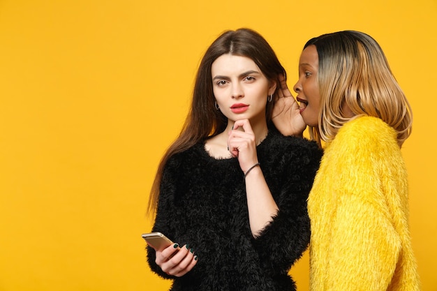 Two young women friends european and african american in black\
yellow clothes standing posing isolated on bright orange wall\
background, studio portrait. people lifestyle concept. mock up copy\
space.