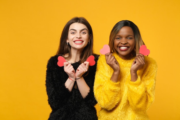 두 젊은 여성 친구 유럽과 아프리카 계 미국인 밝은 주황색 벽 배경, 스튜디오 초상화에 고립 된 포즈 검은 노란색 옷을 입고 서. 사람들이 라이프 스타일 개념입니다. 복사 공간을 비웃습니다.