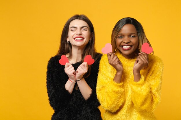 Two young women friends european and african american in black yellow clothes standing posing isolated on bright orange wall background, studio portrait. People lifestyle concept. Mock up copy space.