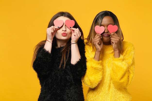 Two young women friends european and african american in black yellow clothes standing posing isolated on bright orange wall background, studio portrait. People lifestyle concept. Mock up copy space.