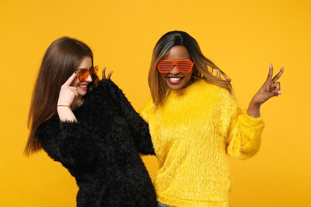 두 젊은 여성 친구 유럽과 아프리카 계 미국인 밝은 주황색 벽 배경, 스튜디오 초상화에 고립 된 포즈 검은 노란색 옷을 입고 서. 사람들이 라이프 스타일 개념입니다. 복사 공간을 비웃습니다.