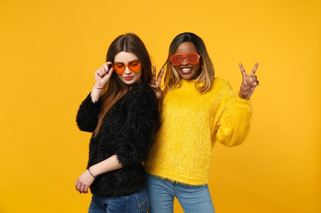 Two young women friends european and african american in black yellow clothes standing posing isolated on bright orange wall background, studio portrait. People lifestyle concept. Mock up copy space.