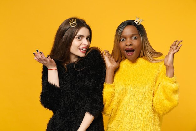 Two young women friends european and african american in black yellow clothes standing posing isolated on bright orange wall background, studio portrait. People lifestyle concept. Mock up copy space.