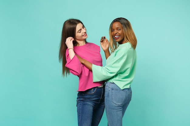 Two young women friends european and african american in pink green clothes dancing fooling isolated on blue turquoise wall background, studio portrait. People lifestyle concept. Mock up copy space.