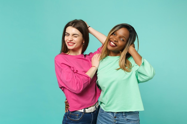 Photo two young women friends european and african american in pink green clothes standing posing isolated on blue turquoise wall background, studio portrait. people lifestyle concept. mock up copy space.