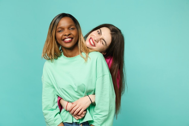 Two young women friends european and african american in pink green clothes standing posing isolated on blue turquoise wall background, studio portrait. People lifestyle concept. Mock up copy space.