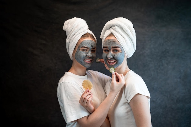 Two young women girlfrined in white towel made beauty clay facial mask and hold orange isolated on black Skin care spa at gome Anti aging treatment