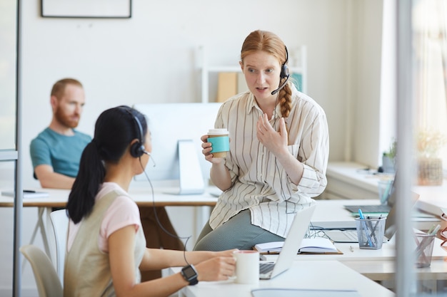 Due giovani donne in cuffia che bevono caffè e parlano tra loro durante la pausa caffè in ufficio