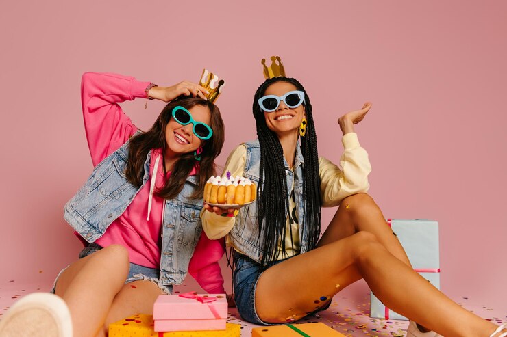 Photo two young women holding birthday cake and smiling while sitting near gift boxes on pink background