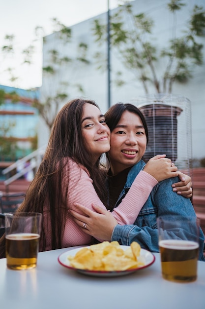 Two young women hugging each other at a bar terrace while\
having fun and looking at camera