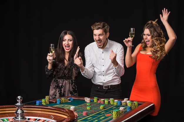 Two young women and man behind roulette table on black background. Emotions players