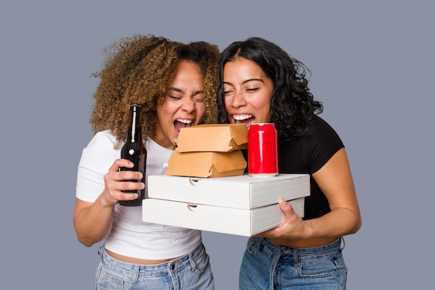 Two young women one Latina and one with Afro hair laugh as they hold pizzas and burgers
