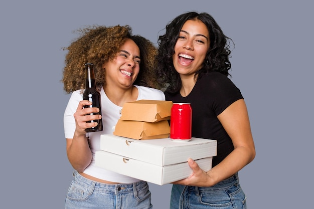 Two young women, one Latina and one with Afro hair laugh as they hold pizzas and burgers