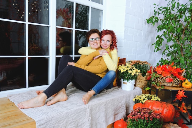 Two young women, sisters, friends in sweaters are sitting in bed at cozy home. Autumn decorations