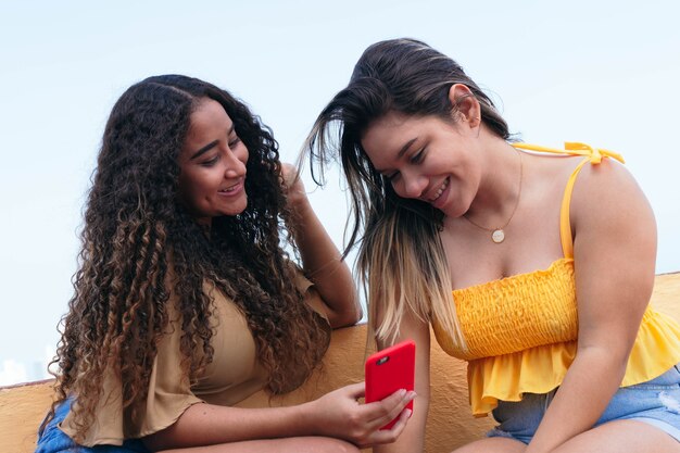 Two young women sitting on city street, looking at cell phone and talking in the street.