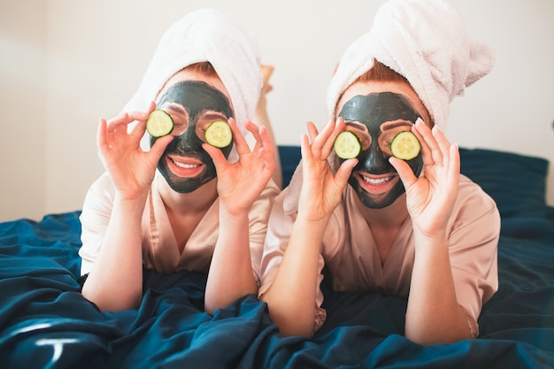 Two young women in towels and pajamas have a fun spa party together in home.