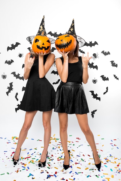 Two young women wearing black dresses, witch hats and high heels hold Halloween pumpkins on their faces .