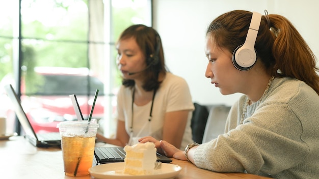 사진 두 명의 젊은 여성이 헤드폰을 착용하고 카페에서 노트북으로 일하고 테이블 위에 케이크와 주스를 고 있습니다.