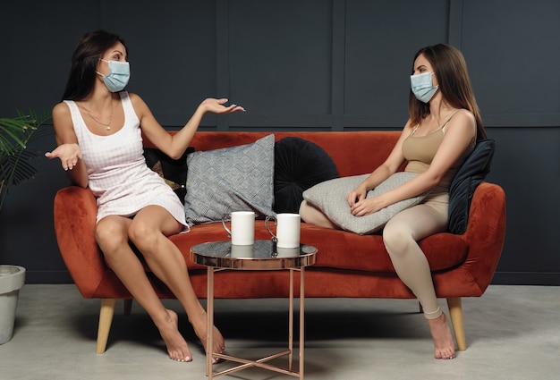 Two young women wearing mask while sitting on couch at home