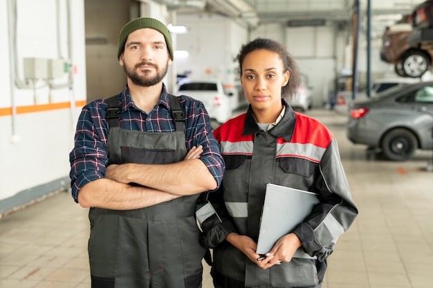 Two young workers of car repair center standing close to each other in front of camera against interior of large workshop with automobiles