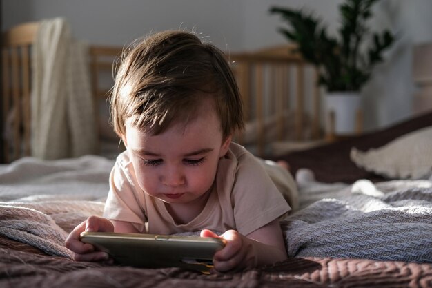 A twoyearold boy lies on a bed and watches cartoons on a smartphone