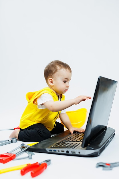 A twoyearold boy in a yellow vest on a white background sits with a laptop and plays with plastic construction tools Toys for small children