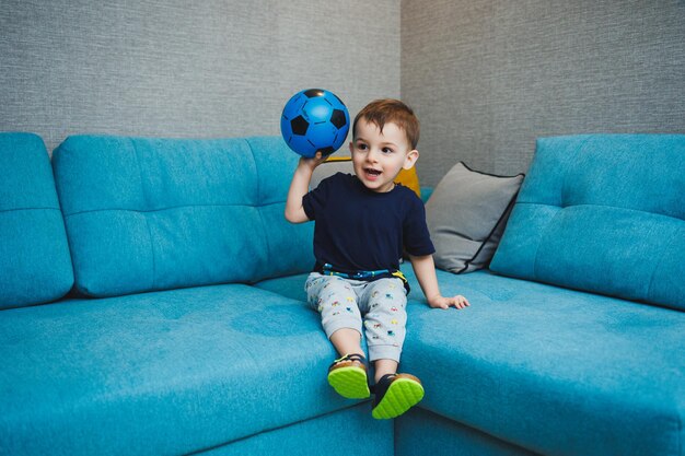 A twoyearold little boy sits on the sofa with a ball in his hands a developed emotional boy plays with a ball at home