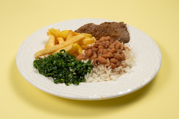 Piatto base tipico brasiliano con riso, fagioli, bistecca, patatine fritte e cavolo brasato in un piatto bianco in vista