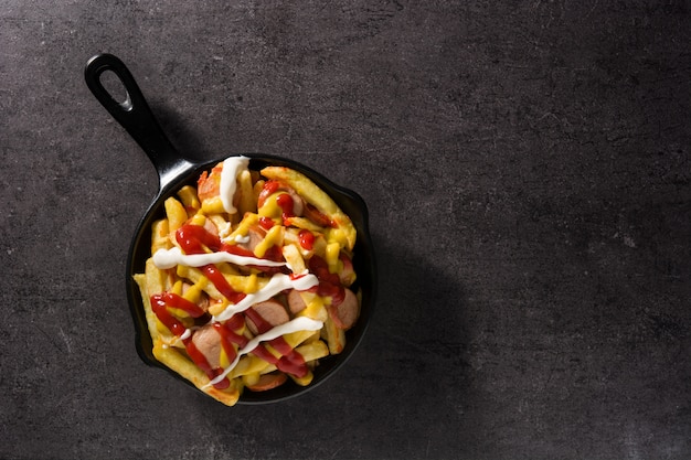 Typical Latin America Salchipapa. Sausages with fries, ketchup, mustard and mayo in iron pan on black background Top view copy space