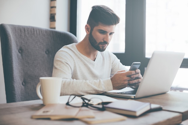 Typing business message. Confident young man holding smart phone and looking at it while sitting at his working place in office