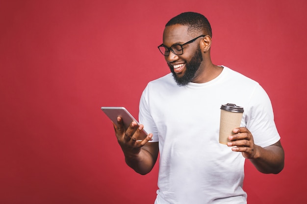 Digitare un messaggio uomo di colore allegro che scrive qualcosa sul telefono cellulare, bevendo caffè e sorridendo mentre stando isolato contro il fondo rosso.