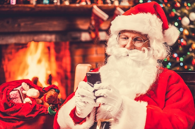 Typing message to elfs. Happy Santa Claus typing a message on the mobile phone and smiling while sitting at his chair with fireplace and Christmas Tree in the background