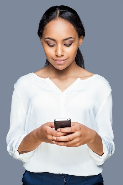 Typing message to friend. Confident young African woman holding mobile phone and looking at it with smile while standing against grey background